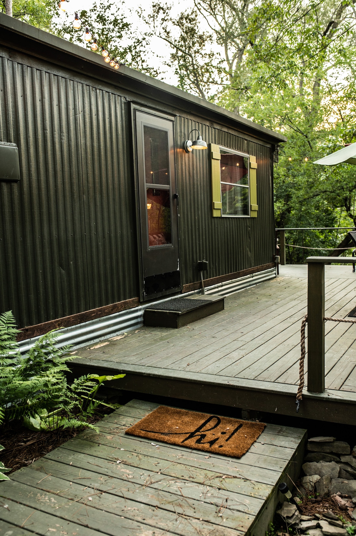 The entrance to the cottage is framed by a wooden deck surrounded by natural greenery. A welcome mat rests at the base of the steps leading up to the door, which features a dark-colored finish and yellow trim. Soft lighting enhances the inviting ambiance.