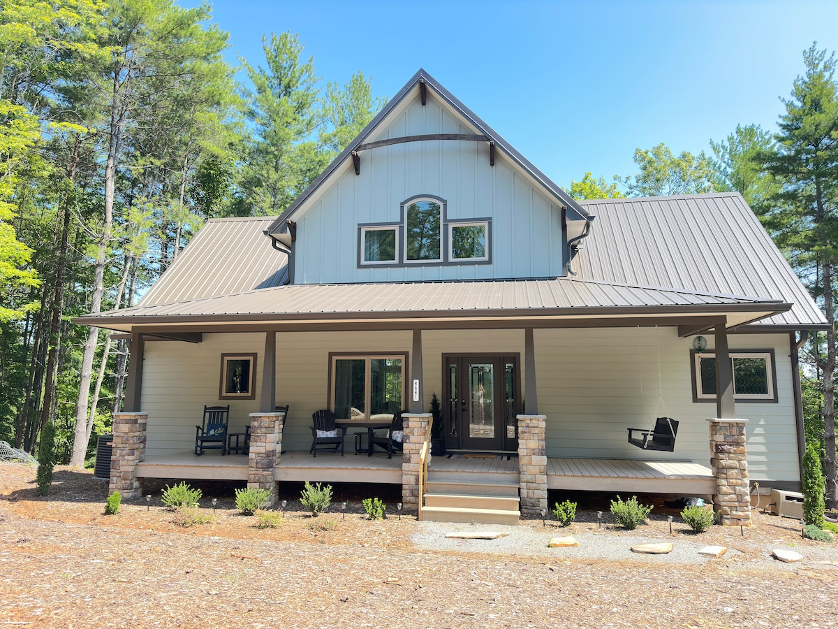 A charming craftsman-style mountain home is presented, featuring a metal roof and a welcoming porch. Stone columns frame the entrance, and seating areas are arranged on the porch. Lush greenery surrounds the property, creating a serene outdoor environment.