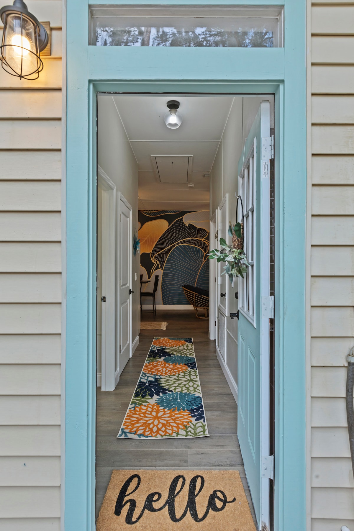 A welcoming entryway is highlighted by a colorful doormat and a vibrant area rug featuring floral patterns. The hallway extends into the distance, where a beautifully designed mural adds a touch of character to the walls. Soft lighting enhances the inviting atmosphere.
