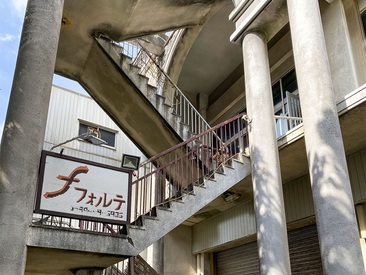 An exterior view of the building features a set of concrete stairs leading to the entrance, bordered by a sturdy railing. The sign of the property is displayed prominently at the base, indicating the name 'フォルテ.' Sunlight casts gentle shadows on the textured surfaces.