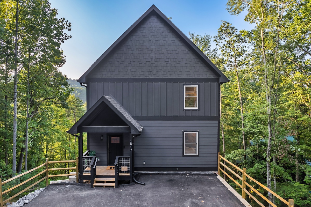The exterior of the cabin features a modern design with dark siding and large windows. An inviting entrance is complemented by a covered porch and steps leading to an asphalt driveway surrounded by natural greenery, offering a peaceful setting in the Smoky Mountains.