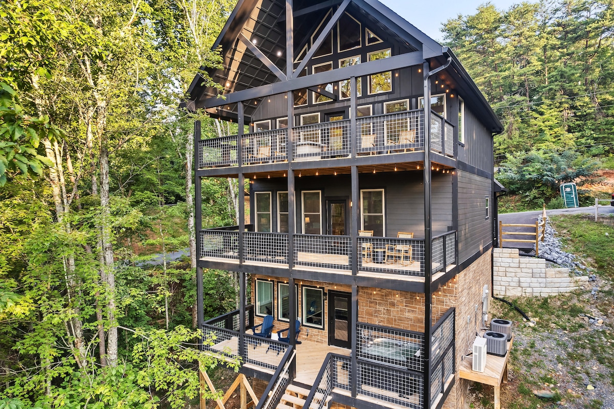 The exterior of the cabin features a modern design with multiple levels and expansive decks. Wood and stone materials create a harmonious blend, while surrounding trees provide a natural backdrop. String lights are visible, hinting at a welcoming outdoor ambiance.