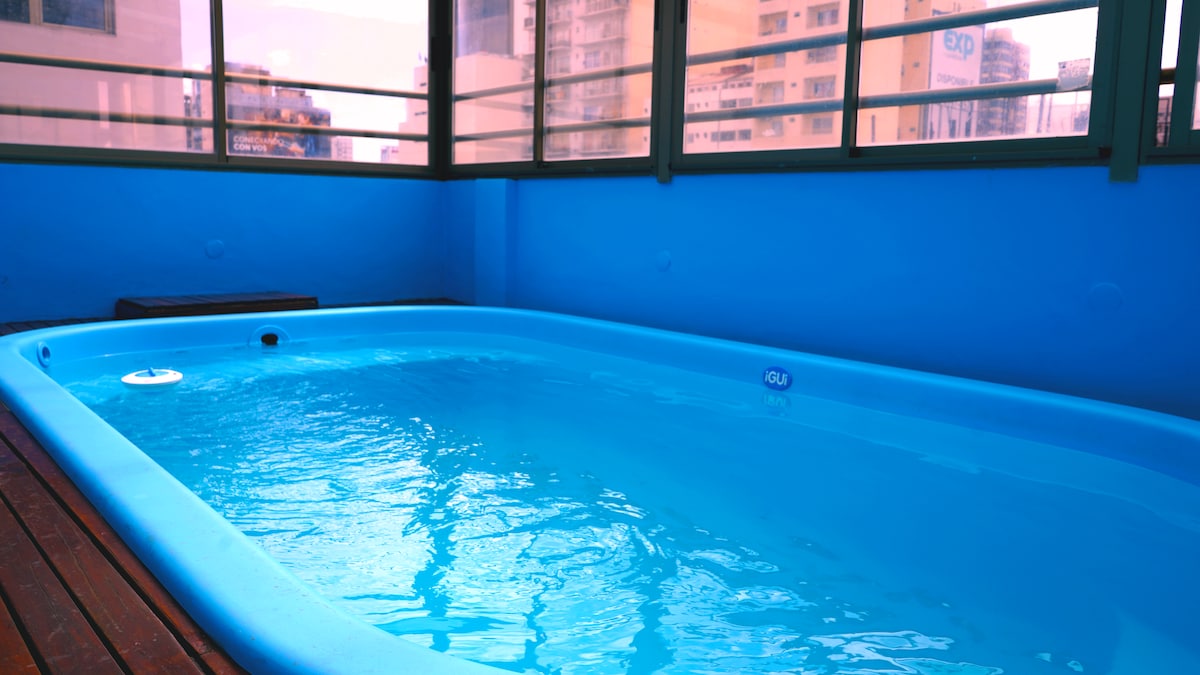 An indoor pool is surrounded by large windows, allowing natural light to fill the space. The water appears clear and inviting, while the walls are painted in a soothing blue hue. Wooden decking complements the tranquil setting.