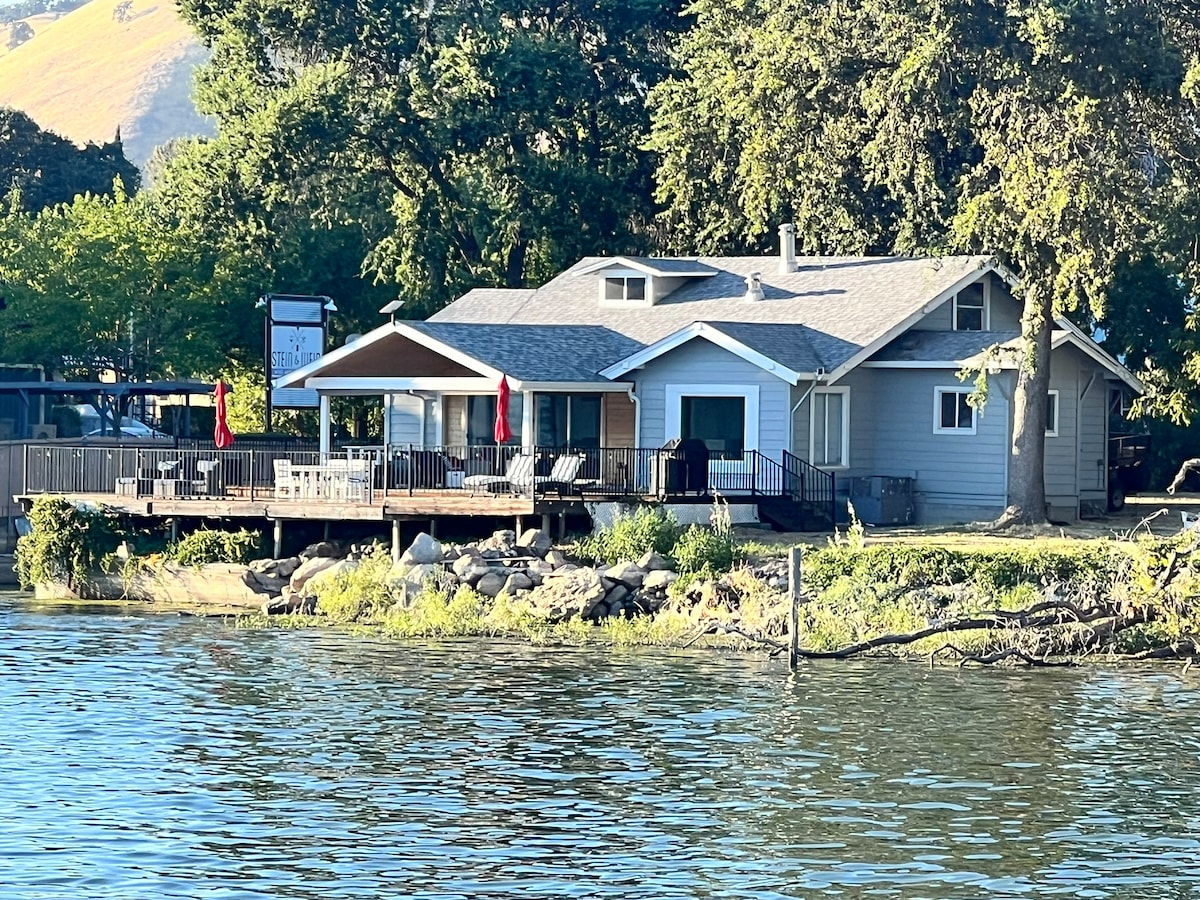 A lakeshore bungalow is viewed from the water, featuring a spacious deck with seating options and a grill. Neatly landscaped areas lead to the home, highlighted by large windows and a welcoming patio. Red umbrellas add a pop of color against the serene backdrop of greenery.