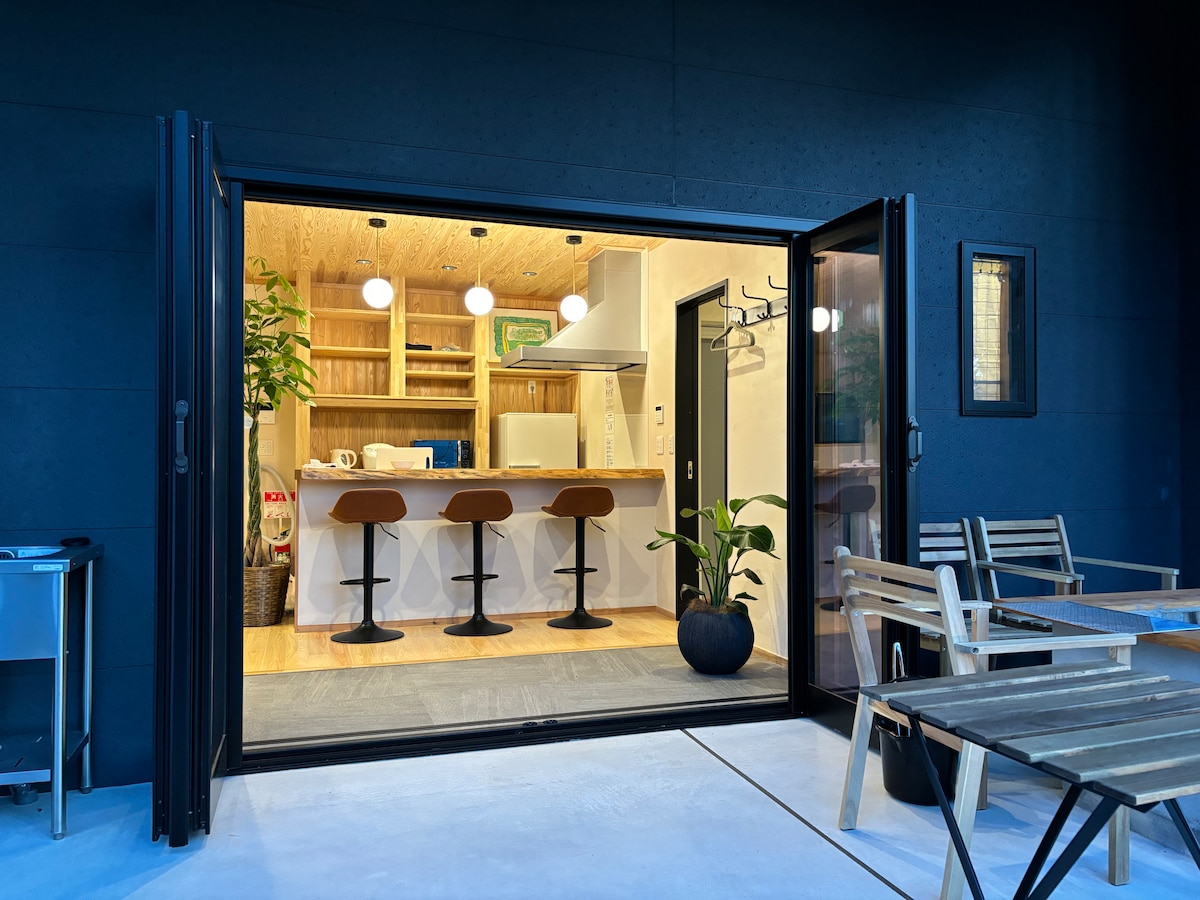 A modern kitchen space is visible through open folding doors, featuring wooden shelving and three bar stools positioned at a counter. A potted plant adds a touch of greenery, while outdoor seating is arranged on the wooden terrace.
