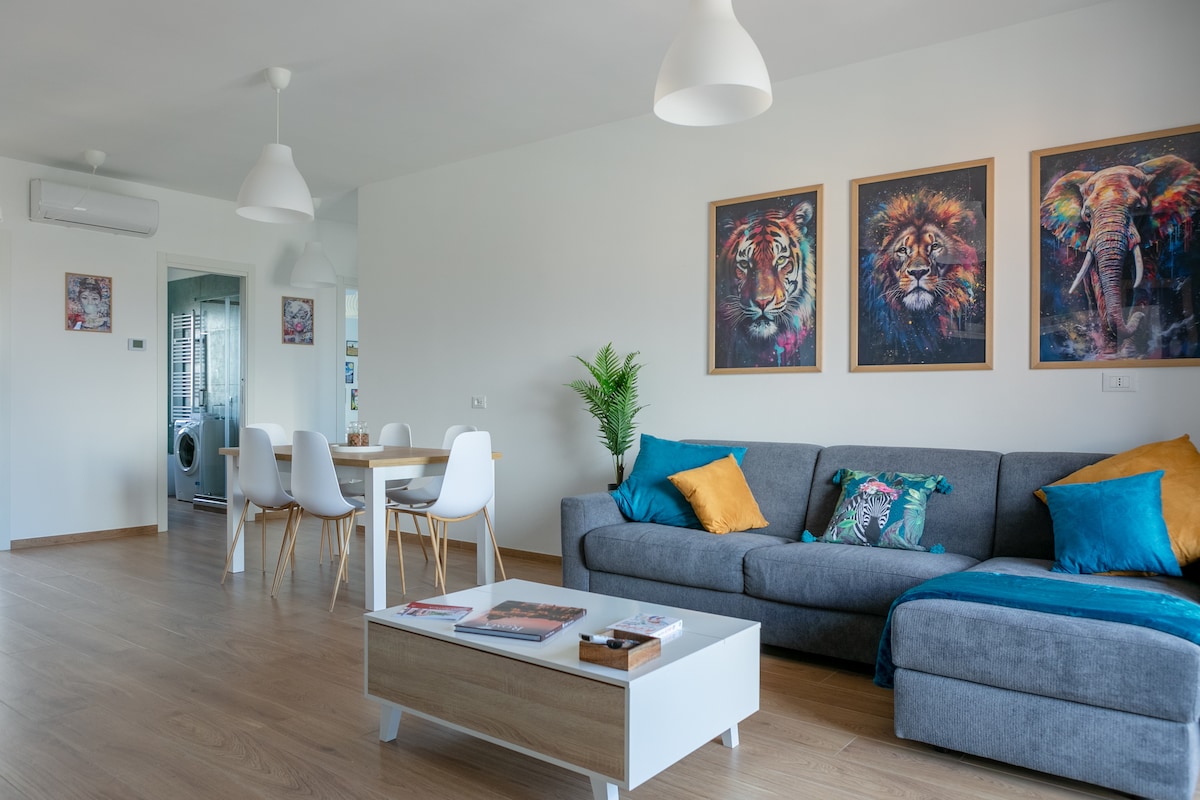 A modern living area showcases a gray sofa adorned with colorful cushions. Three framed animal portraits add interest to the white walls. A dining table with white chairs is positioned in the background, and natural light fills the space through the window.