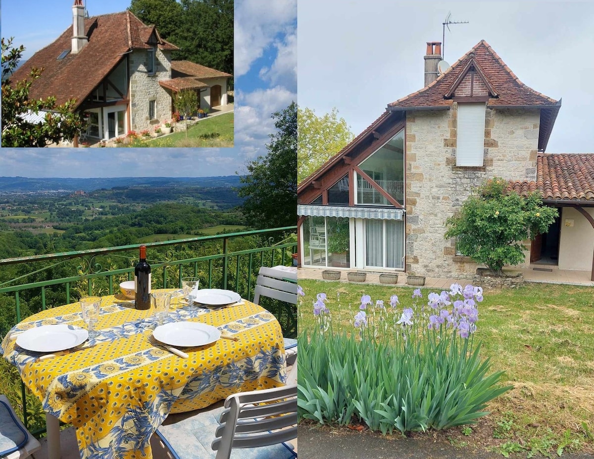 The image features a charming two-story house with a sloped roof and large windows, offering expansive views of the Dordogne valley. A dining table is set outside on a terrace, adorned with a yellow tablecloth, plates, and a bottle of wine. Lavish greenery and blooming flowers complement the outdoor space.