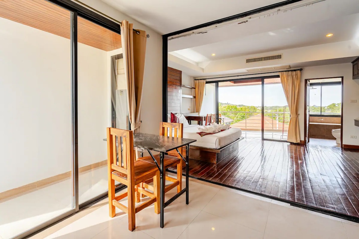 A bright and airy apartment space features large glass doors allowing natural light to flood the room. A wooden dining table with two chairs is positioned near the entrance. In the background, a spacious bed complements the scenic views visible through the sliding doors.