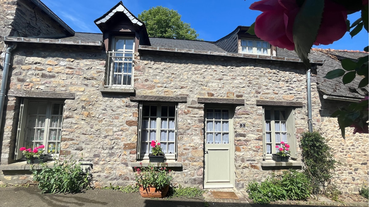 A charming stone house presents a traditional facade, featuring multiple windows adorned with flower boxes filled with bright blooms. Sunlight illuminates the exterior, highlighting the texture of the stone. A well-kept path leads to the entrance, framed by greenery and vibrant flower arrangements.