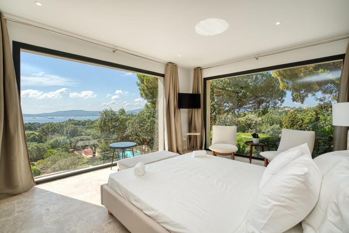 A spacious bedroom is presented with large windows offering panoramic sea views. The room features a queen-size bed with white linens. Two accent chairs and a small table provide a relaxing seating area, while natural light enhances the serene ambiance.