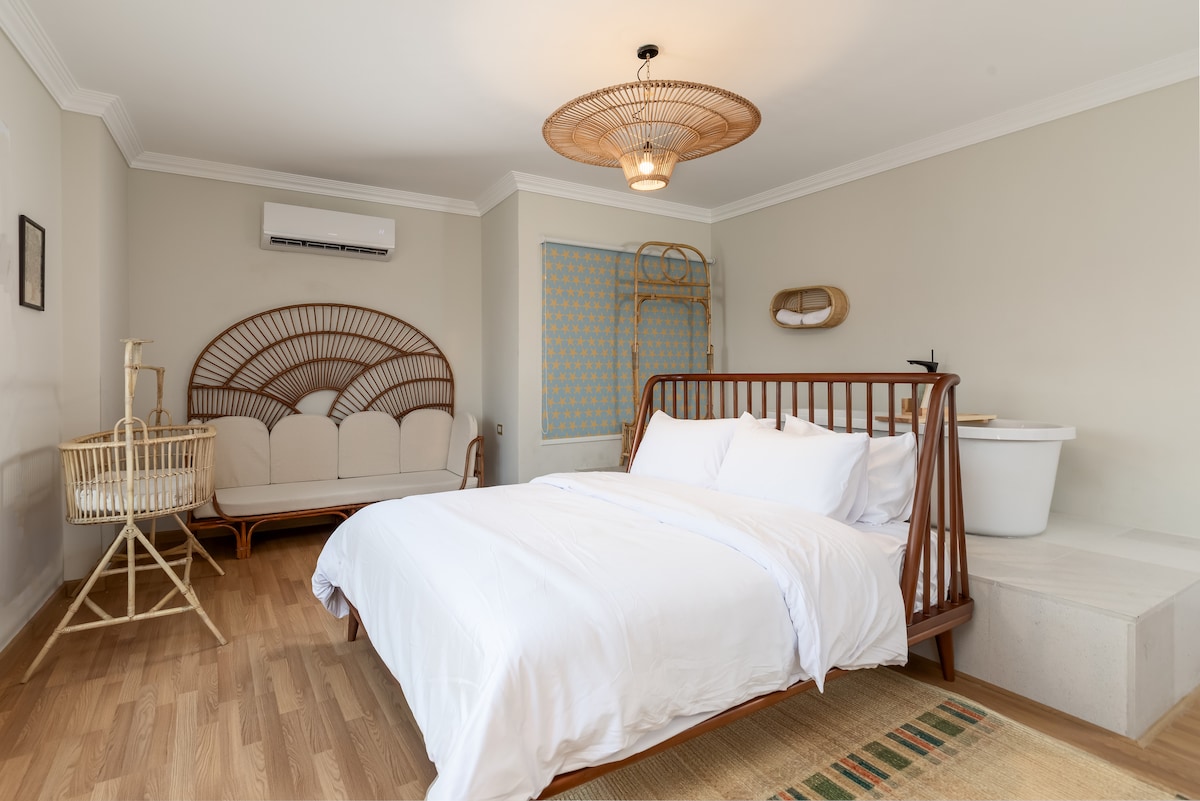 A cozy sleeping area features a spacious bed dressed in crisp white linens. A light hardwood floor complements the neutral wall tones. A wicker accent chair and a rattan seating area add texture, while a soft rug creates a welcoming atmosphere.