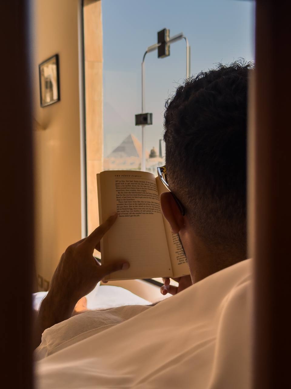 A person is seen reading a book while sitting comfortably in bed. The view through a window features the iconic silhouette of a pyramid in the distance, enhancing the serene atmosphere of the space.