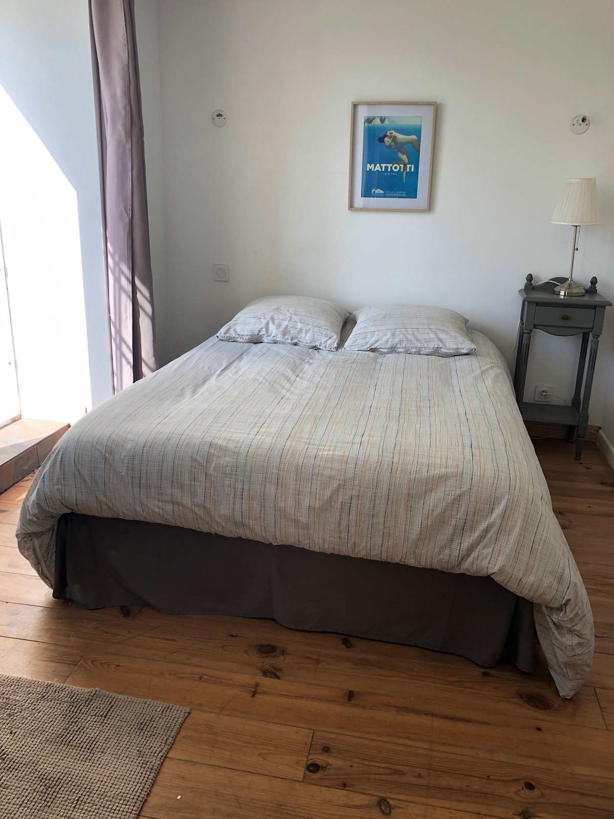 A spacious bed with a light-colored striped duvet is positioned against a wall. Natural light filters in from a nearby window, illuminating the wooden floor. A small bedside table holds a lamp, and a framed artwork is mounted on the wall.
