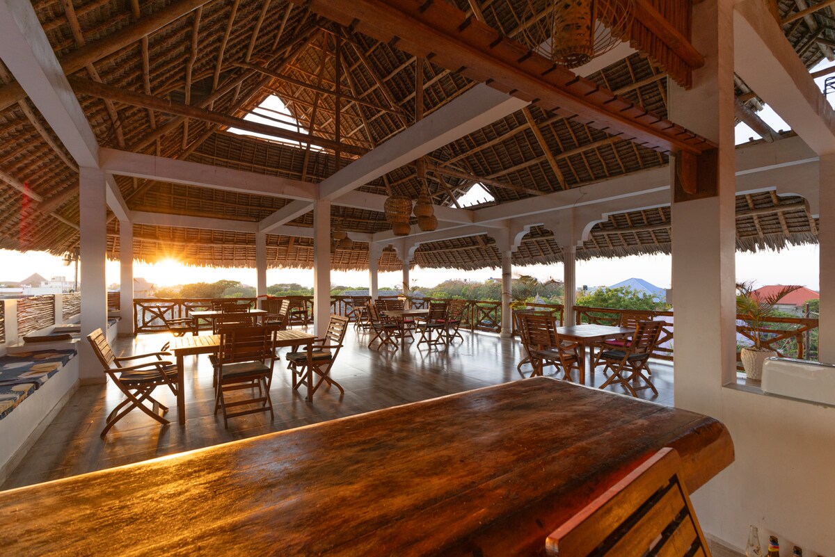 An open dining area is featured with wooden tables and chairs arranged across a spacious floor. High ceilings with thatched roofing enhance airflow. Warm light from the setting sun creates a relaxed ambiance, illuminating the surrounding greenery visible through archways.