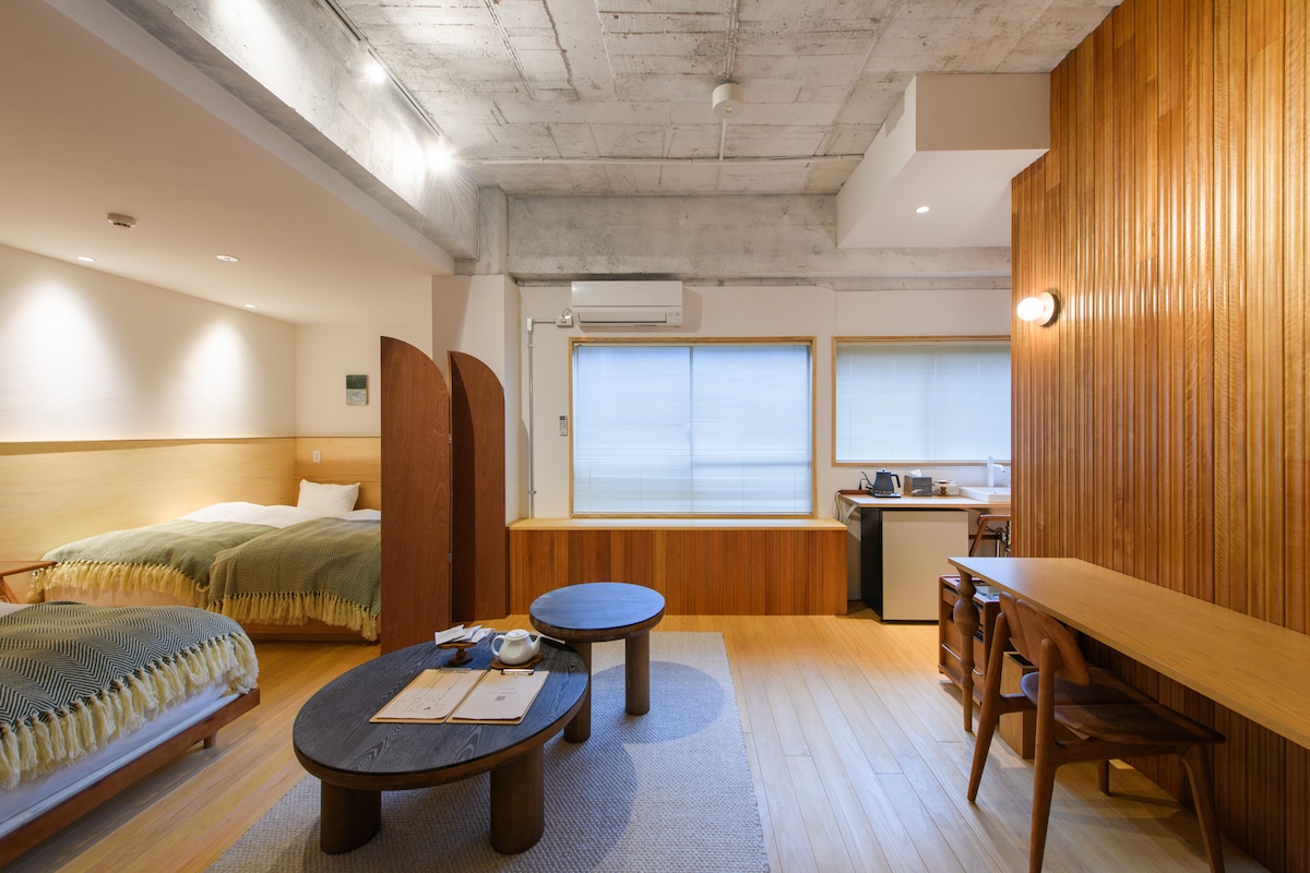 A spacious room features two single beds with textured green blankets, complemented by warm wooden accents. A low circular table sits at the center, surrounded by chairs. Soft light enters through large windows, highlighting the functional kitchenette and work desk area.