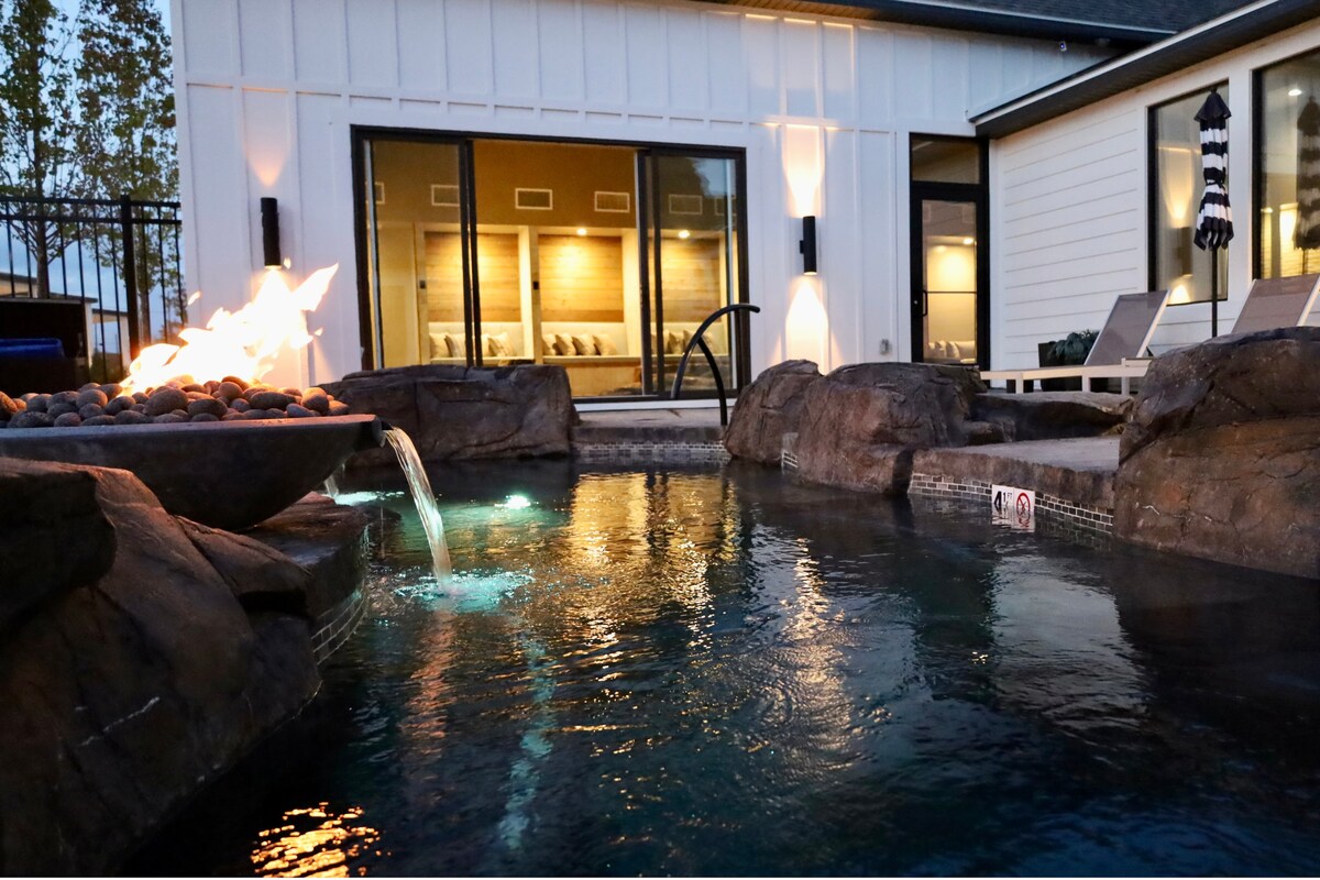 A tranquil hot pool is surrounded by natural stone features and illuminated water jets. A fire pit glows warmly nearby, casting a soft light on the adjacent cottage, which features large glass doors revealing a cozy interior.