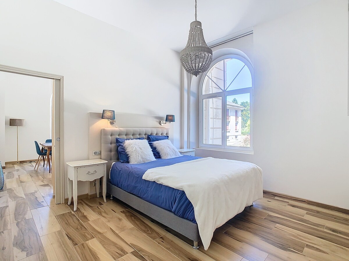 A spacious bedroom features a comfortable bed dressed in soft linens, flanked by stylish bedside lamps. Natural light floods the room through a large arched window. Light wood flooring complements the decor, enhancing the calming atmosphere of the space.