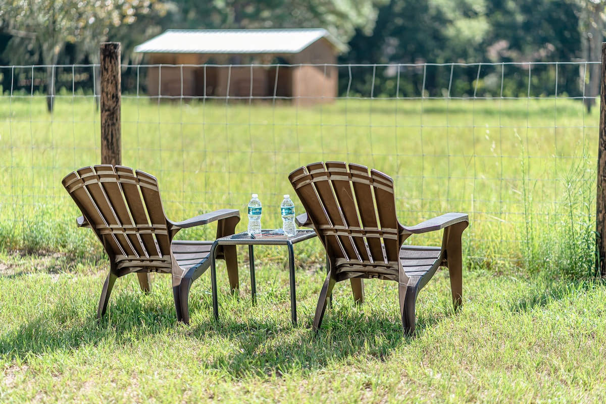 Two wooden chairs and a small table are positioned on grass, overlooking a fenced field. Bottled water sits on the table, and a barn is visible in the background, providing a serene countryside setting.