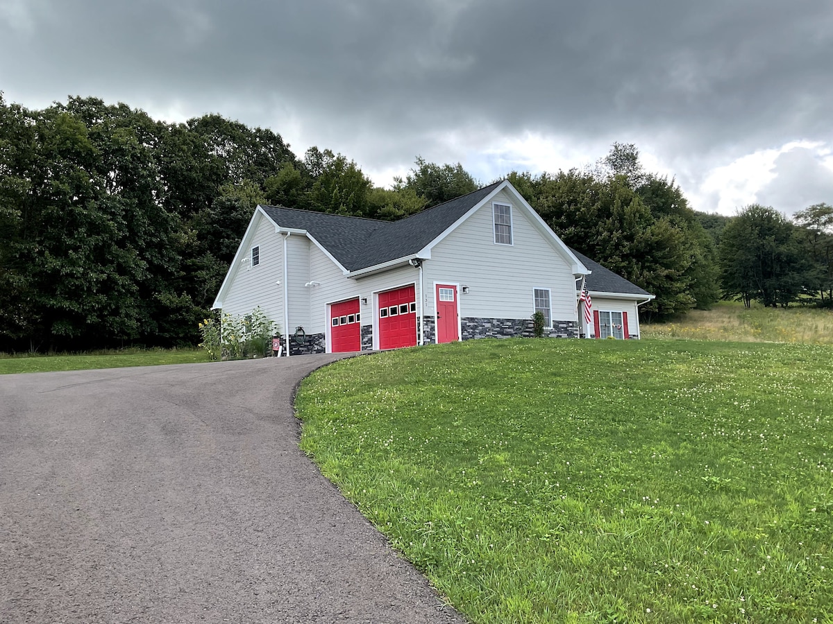 A modern two-story home is set against a backdrop of greenery, featuring a smooth driveway leading to the entrance. The exterior is highlighted by red garage doors and a mix of siding and stone accents, showcasing a balanced design harmonizing with the natural surroundings.