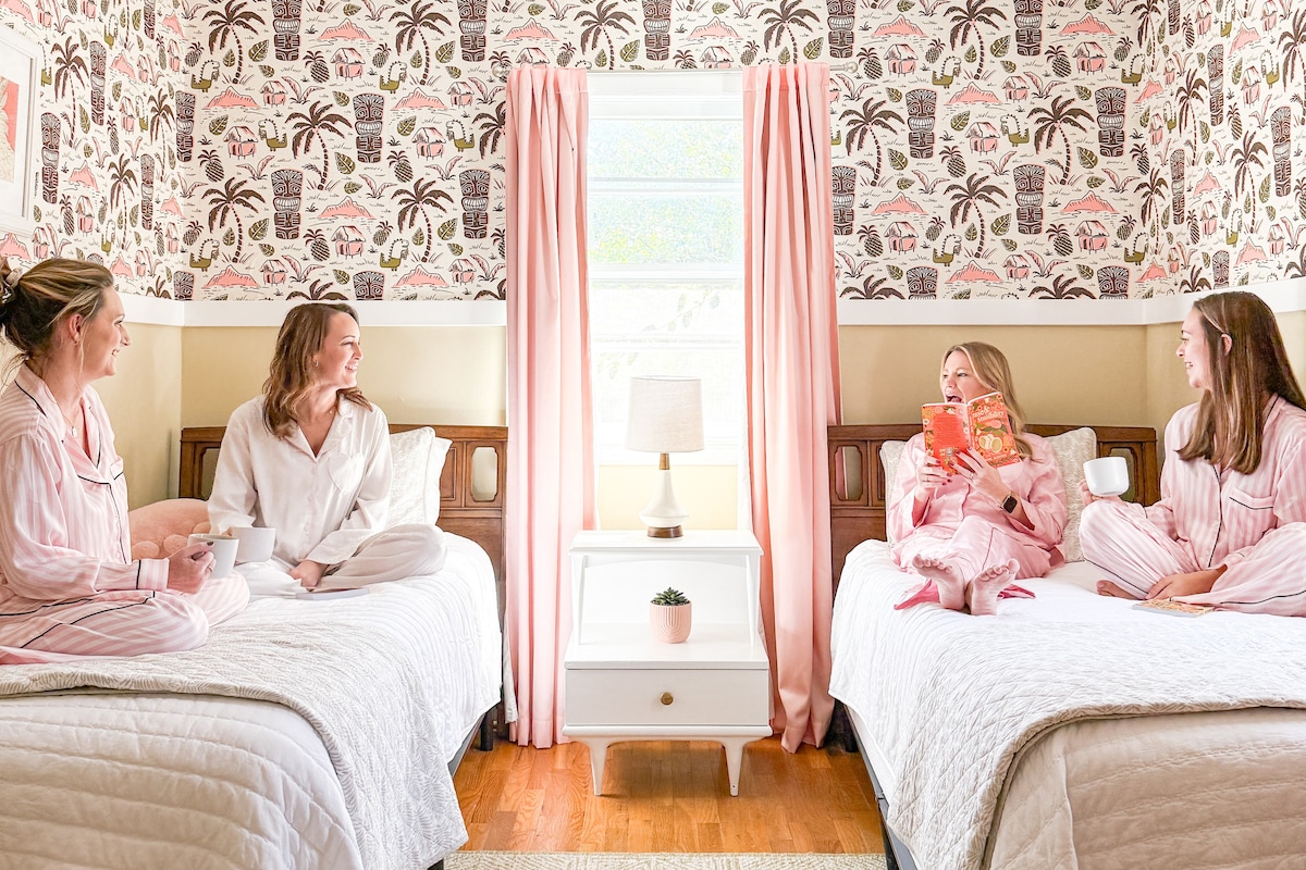 Two cozy twin beds are positioned on either side of a bright window, dressed in light linens. A small bedside table with a lamp sits between the beds. Colorful wallpaper featuring tropical patterns enhances the cheerful atmosphere, while pink curtains frame the window.