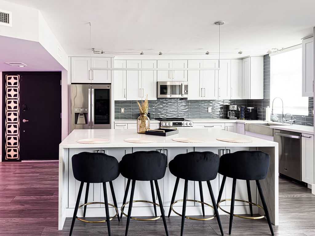 A modern kitchen features sleek white cabinetry and a large island with black bar stools. Upgraded appliances, including a refrigerator and microwave, are visible. The backsplash displays a stylish tile pattern, and natural light streams through windows, creating a bright atmosphere.