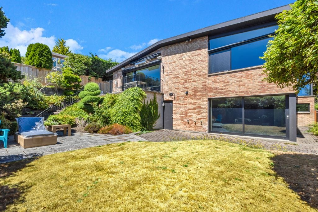 The exterior of a modern home is displayed, featuring large glass windows and brick walls. A spacious, landscaped backyard is visible, complemented by a stone patio and well-maintained greenery. Sunlight illuminates the area, creating a bright atmosphere for outdoor relaxation.