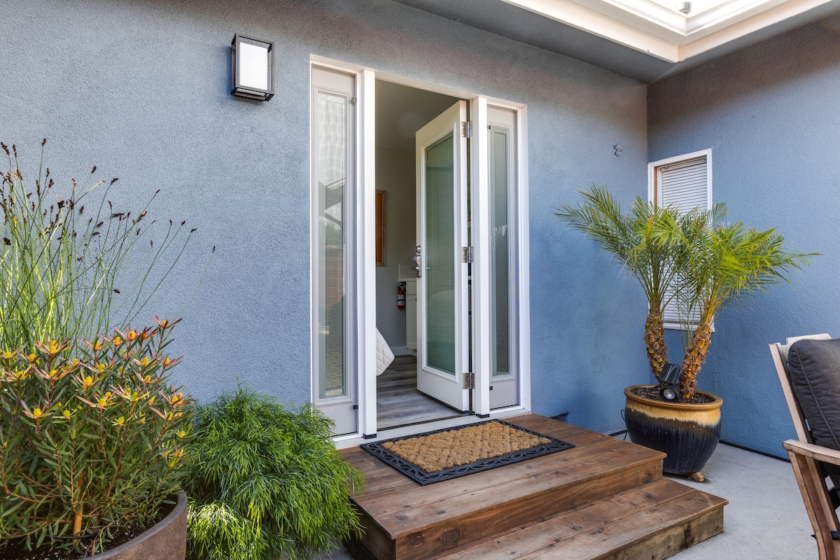 A well-lit entrance features double glass doors that open onto a wooden deck. Lush green plants in decorative pots enhance the inviting entryway, while a doormat welcomes guests. The exterior wall is painted in a calming blue, adding to the overall serenity of the space.