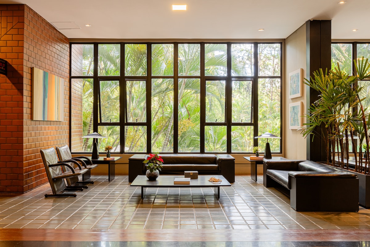 The lobby area features large windows that allow natural light to fill the space, alongside views of lush greenery. Comfortable seating options include dark leather sofas and armchairs. A coffee table with decorative items sits at the center, creating a welcoming atmosphere.