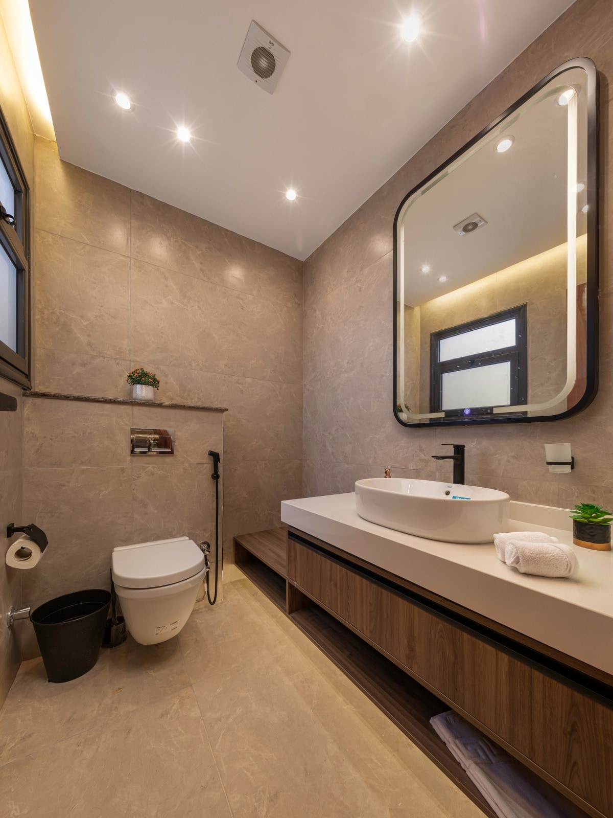 A modern bathroom is featured, showcasing stylish finishes. A large mirror is mounted above a sleek sink, paired with a wooden storage unit. A toilet and a small waste bin are present, along with a touch of greenery from a small plant on the counter.