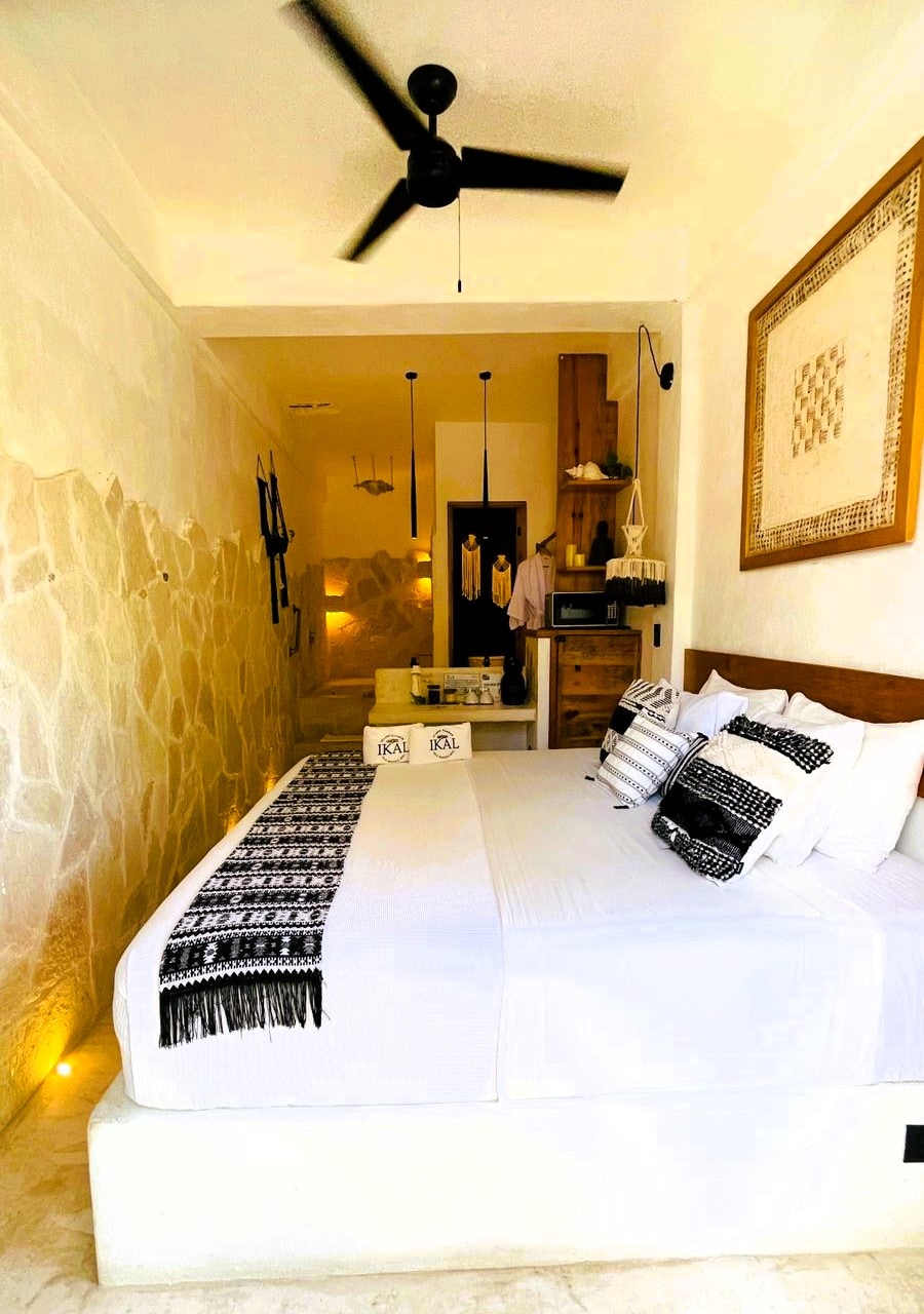 A cozy bedroom features a spacious bed adorned with a textured black and white throw. Natural stone walls provide a rustic backdrop, while ambient lighting creates a soft glow. A ceiling fan is visible, and built-in storage elements contribute to the room's functionality.