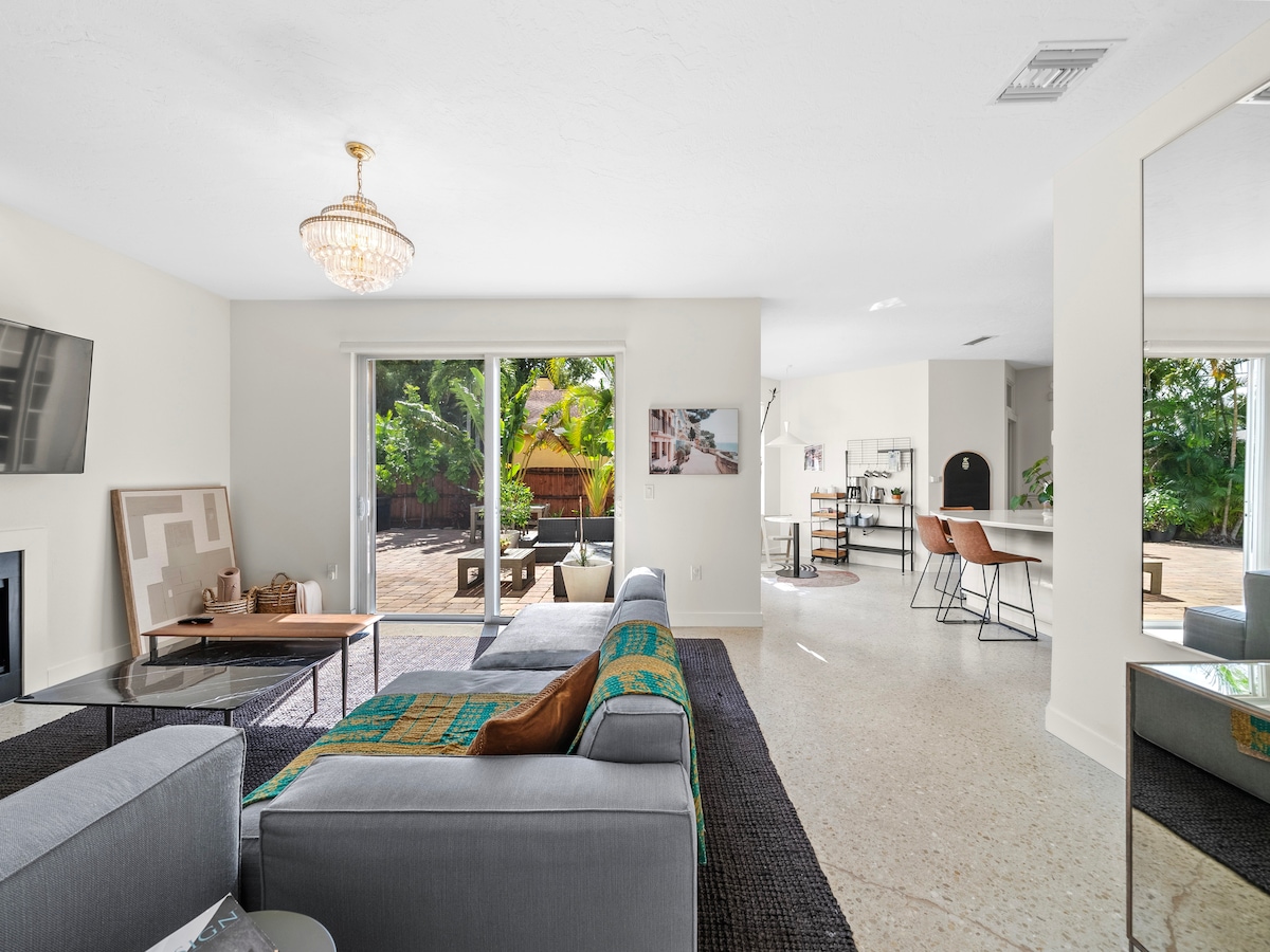 A spacious living area features a comfortable gray sofa adorned with a colorful throw. Large glass doors provide access to the patio, surrounded by lush greenery. A modern chandelier hangs from the ceiling, while a mirror reflects natural light and the open kitchen area in the background.