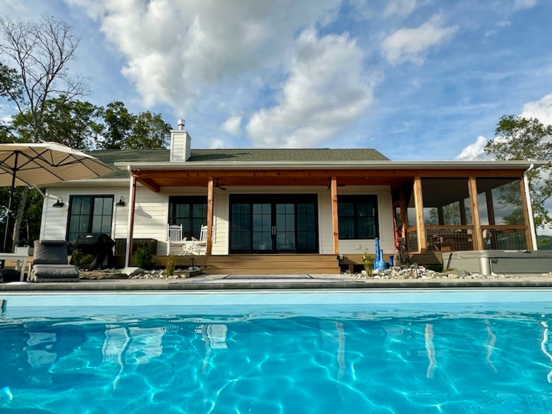 The exterior of the house showcases a modern design with large glass doors opening to a spacious patio. The inviting pool reflects the vibrant sky, while a canopy provides shade. The surrounding area is landscaped with pebbles, enhancing the outdoor space.