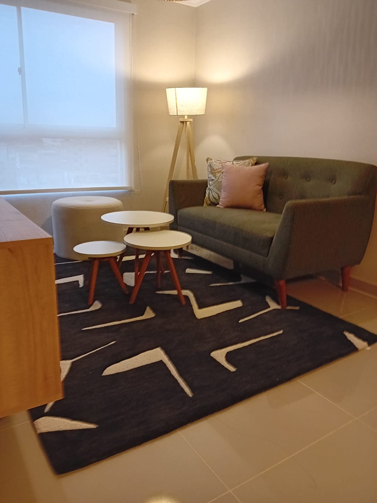 A cozy seating area is featured, highlighted by a green armchair with wooden legs and a small cushion. Two minimalist white side tables are placed on a patterned dark rug, with a standing lamp illuminating the space. A large window provides natural light.