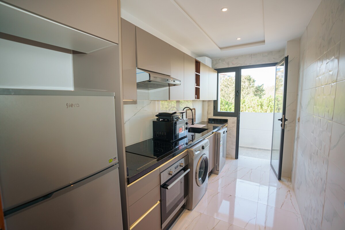A modern kitchen is equipped with sleek cabinetry and a black countertop. A silver refrigerator, washing machine, and various appliances are visible. Natural light enters from a window, highlighting the spacious layout and providing a view of the outside greenery.