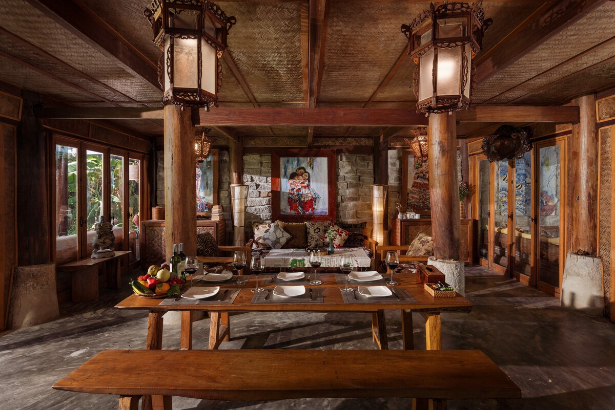A spacious dining area features a long wooden table set with dishes and glasses. Natural light enters through large windows, illuminating the rustic wooden beams and columns. Artwork depicting local culture adorns the walls, enhancing the area’s inviting ambiance.