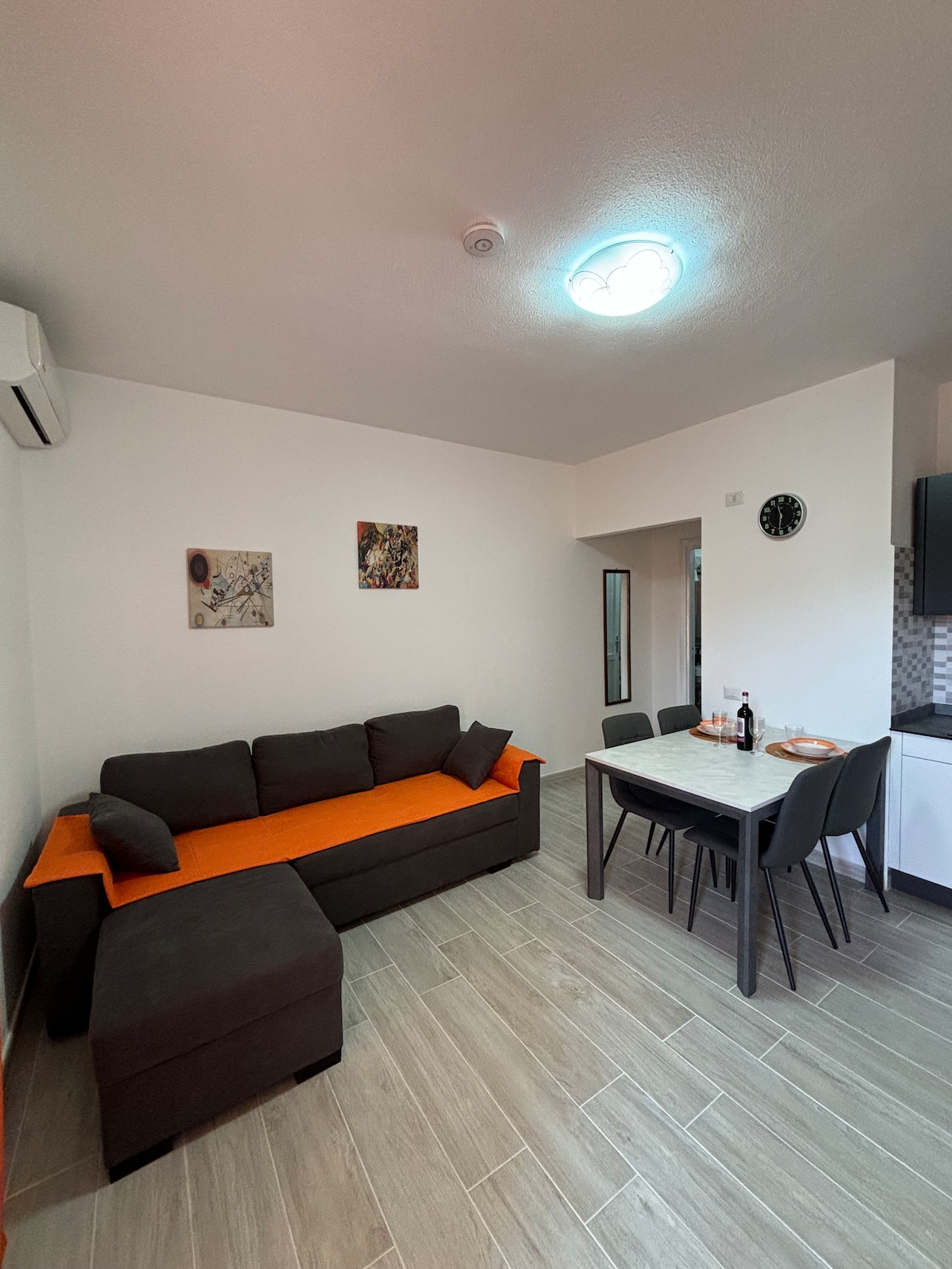 A modern living area is shown, featuring a comfortable couch with an orange accent, a dining table with chairs, and an air conditioning unit mounted on the wall. Colorful artwork is displayed on the white walls, and natural light illuminates the light-colored flooring.