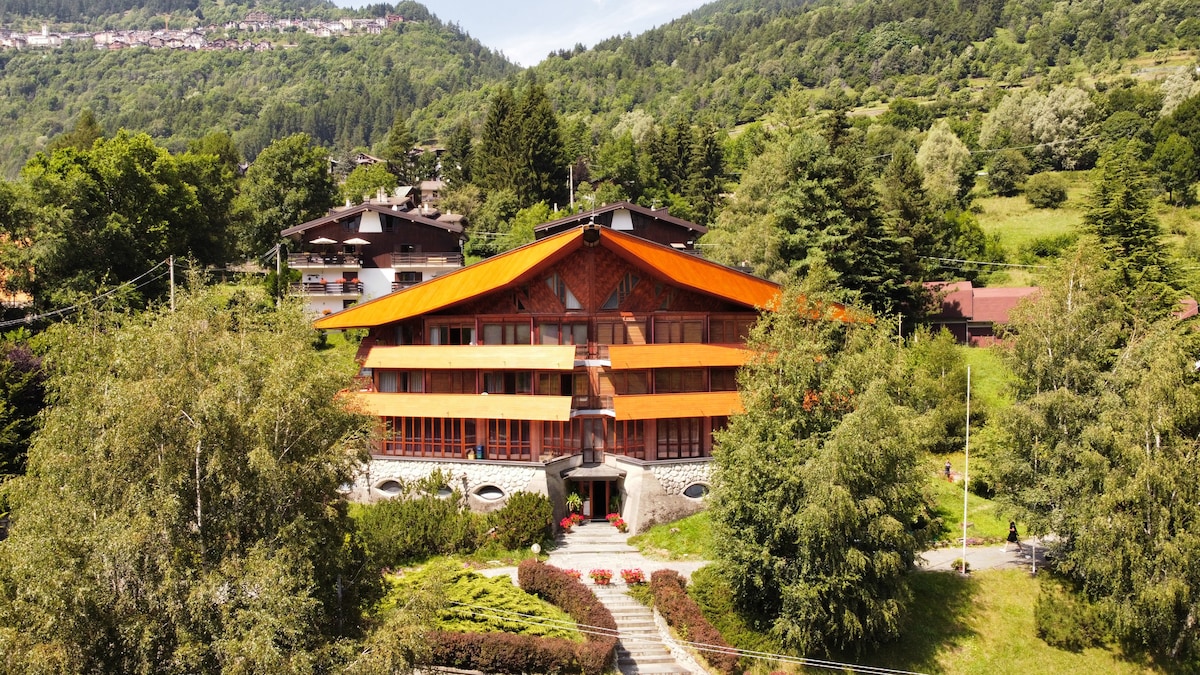 The wooden hotel features a striking orange roof and multiple balconies, set against a lush mountain backdrop. Surrounding greenery and well-maintained pathways highlight the property's outdoor space, with nearby trees providing additional shade and natural beauty.