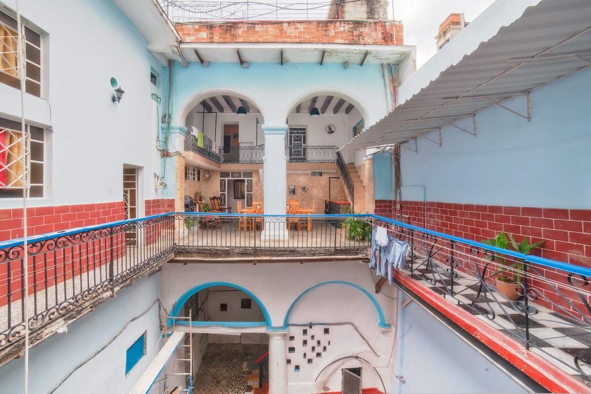 An open-air courtyard is surrounded by colorful walls and balconies with wrought-iron railings. Potted plants and furniture are visible on the balconies. Within the courtyard, a patterned floor contrasts with the blue and red accents, creating a lively communal space.