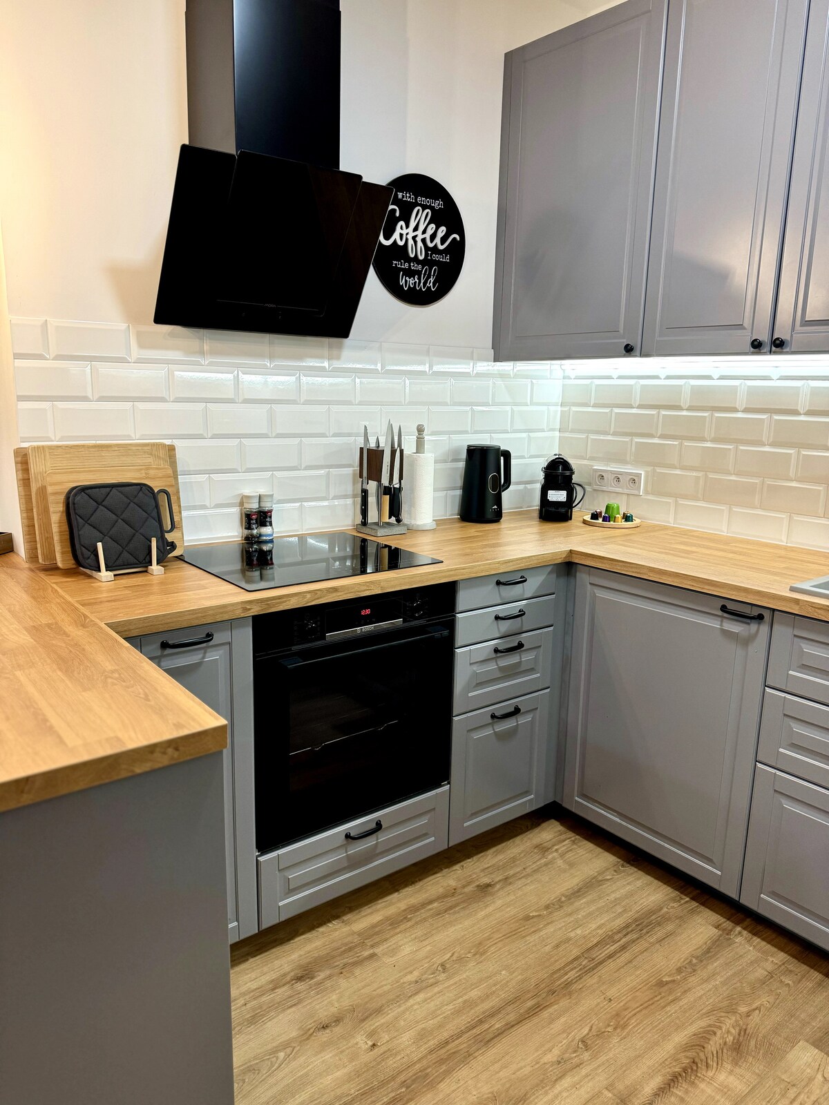 A modern kitchen is highlighted by gray cabinetry and a wooden countertop. Appliances include an oven, a stovetop, and a coffee maker. Neutral tiles create a clean backdrop, while kitchen utensils are organized neatly. The space is well-lit, offering a functional and inviting cooking area.