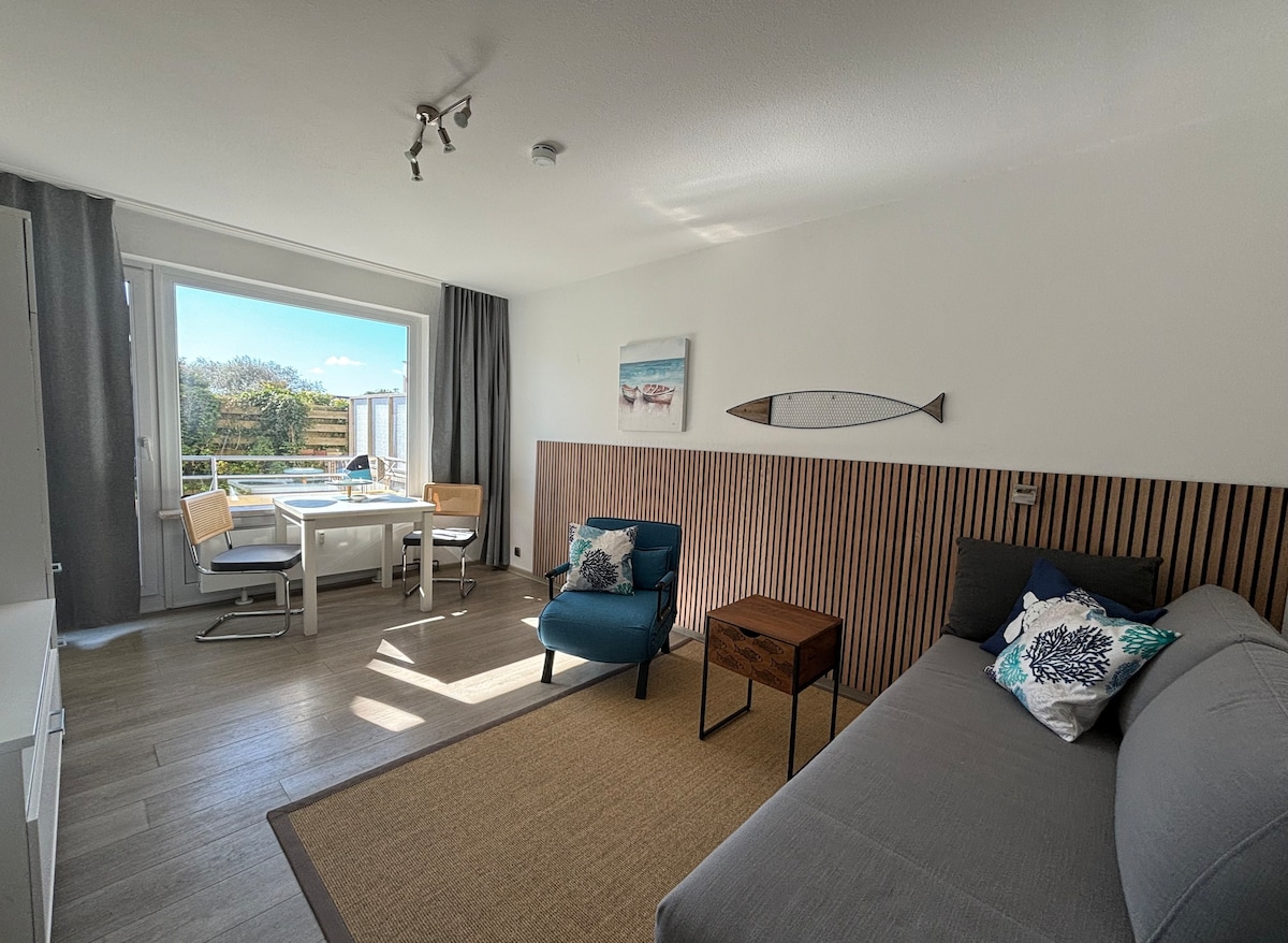 A light-filled living area features a cozy seating arrangement with a gray sofa bed and a blue chair. A small wooden table is positioned beside it. Large glass doors open to a balcony, providing scenic views and natural light. Neutral tones and wood accents create a harmonious space.