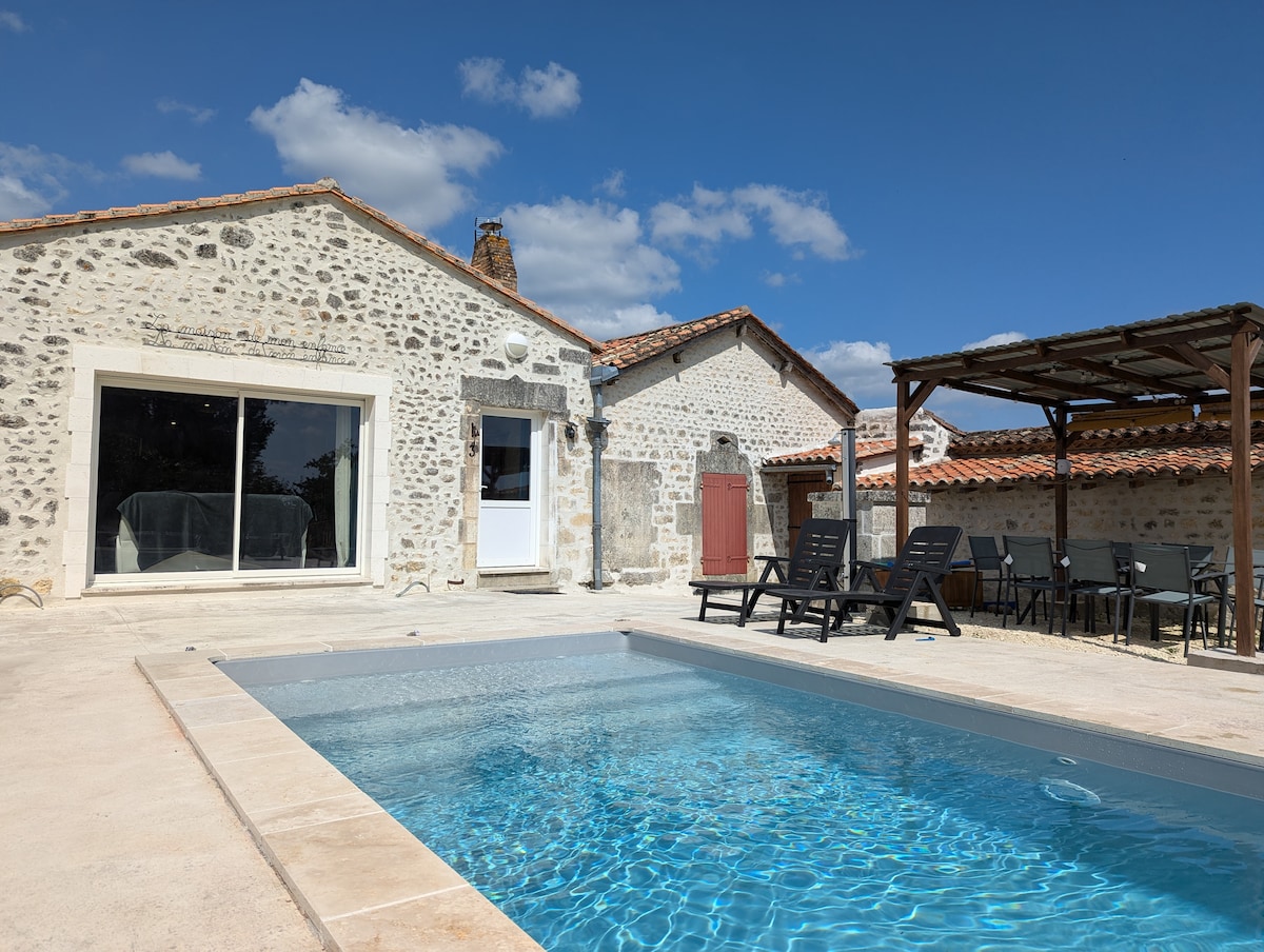 The exterior of a charming stone house features a private swimming pool surrounded by a spacious terrace. A shaded dining area is present nearby, and several lounge chairs are positioned around the pool. Clear blue skies enhance the inviting ambiance of the outdoor space.