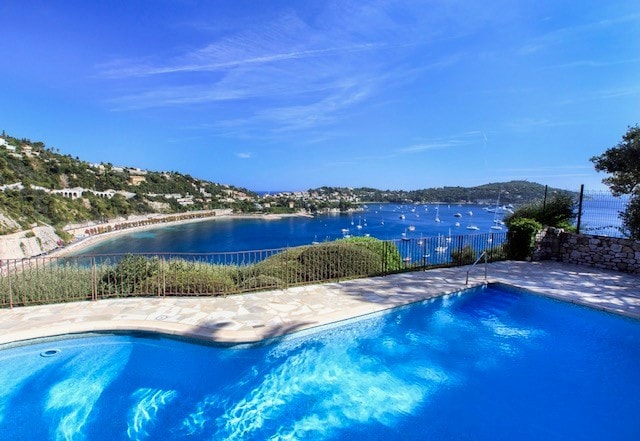An inviting swimming pool is framed by a stone patio, offering scenic views of the calm blue waters and sailboats in the distance. Lush greenery and gentle hills surround the waterfront, under a clear blue sky.