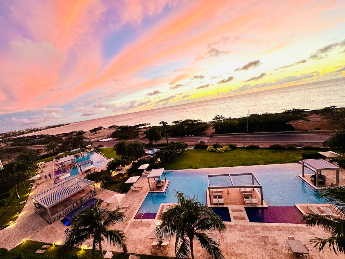 The image captures a serene view from a fifth-floor balcony, showing an infinity pool surrounded by palm trees. A pastel sunset paints the sky above the turquoise Caribbean waters, emphasizing the peacefulness of the resort area below.