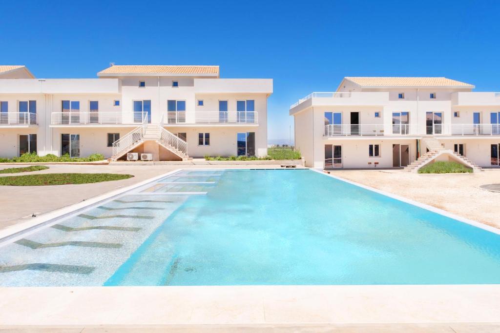 A spacious outdoor area features a large, inviting swimming pool bordered by modern buildings. Sunlight reflects off the clear water, creating a bright ambiance. Lush greenery surrounds the space, enhancing the serene environment.