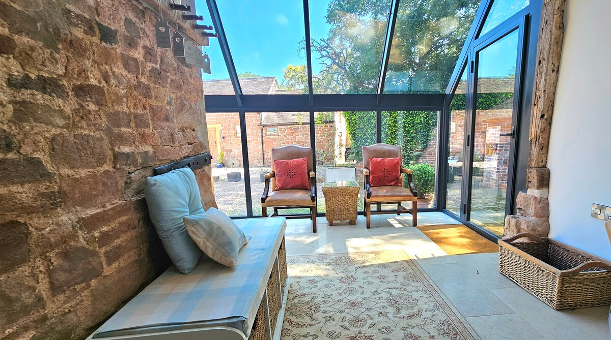 The atrium is designed with glass panels, allowing natural light to fill the space. Comfortable chairs are positioned around a small table, situated on a tiled floor. A decorative area rug adds warmth, while views of the enclosed garden are visible through the sliding glass doors.
