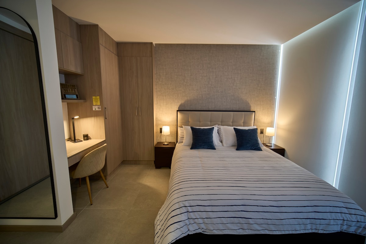 A well-appointed sleeping area features a spacious bed adorned with striped linens and two blue accent pillows. Soft lighting is provided by two bedside lamps, enhancing the serene atmosphere. A functional workspace with a desk and a stylish chair is positioned nearby, complemented by a built-in mirror and ample storage.