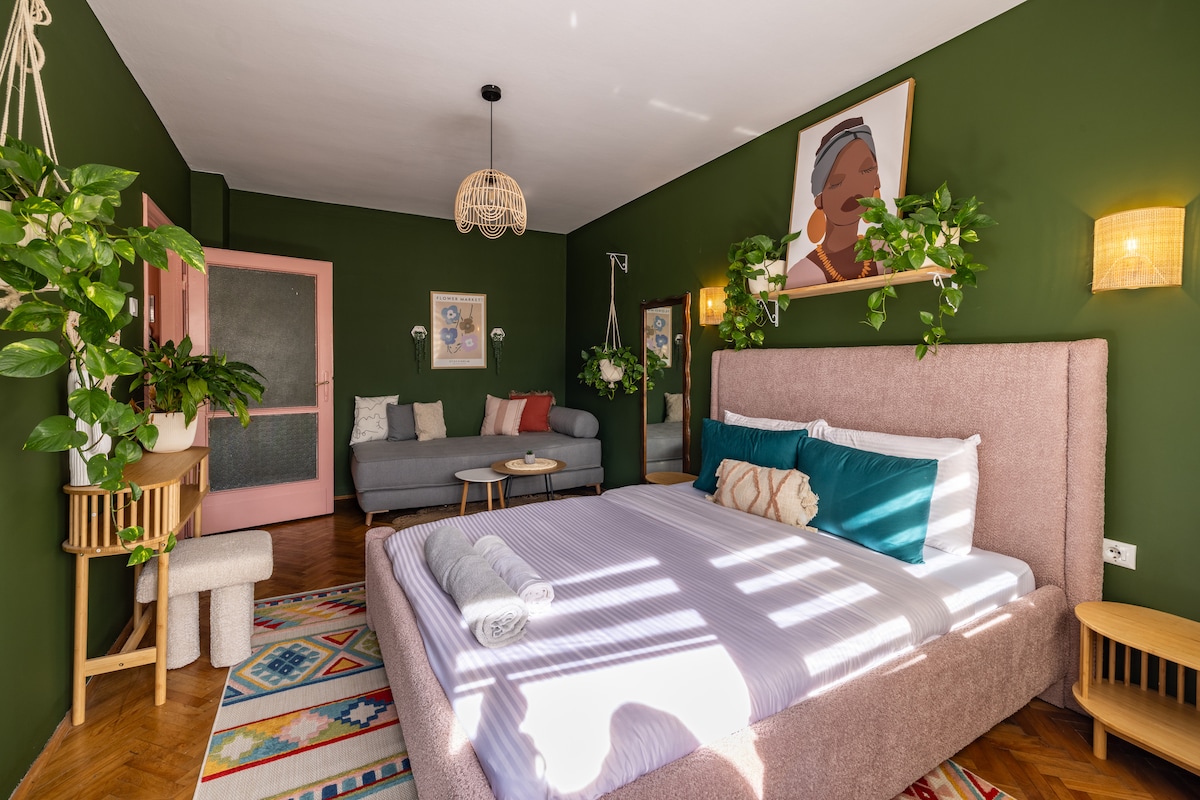 The bedroom features a spacious bed with soft linens and colorful pillows, surrounded by vibrant green walls. A seating area with a gray sofa and a small table is present. Wall-mounted plants and lamps provide subtle lighting, while a colorful rug adds texture to the floor.