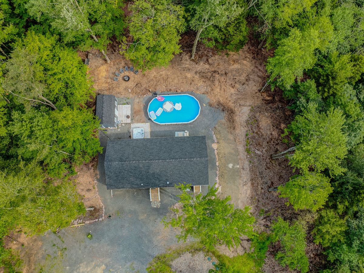 An aerial view of the retreat is presented, highlighting the private pool surrounded by lounge chairs, nestled within lush greenery. The outdoor fire pit is visible, along with the spacious structure of the retreat, blending harmoniously with the natural landscape.