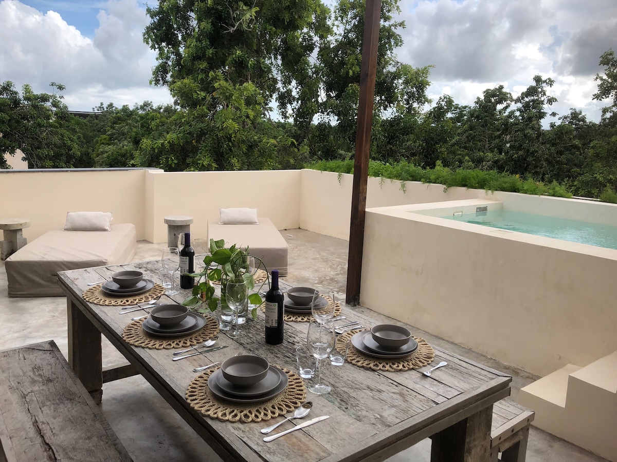 An outdoor dining table is set with plates, glasses, and wine bottles, placed on a textured wooden surface. Soft lounge chairs are situated nearby under a canopy of lush greenery, with a private pool visible in the background against a cloudy sky.