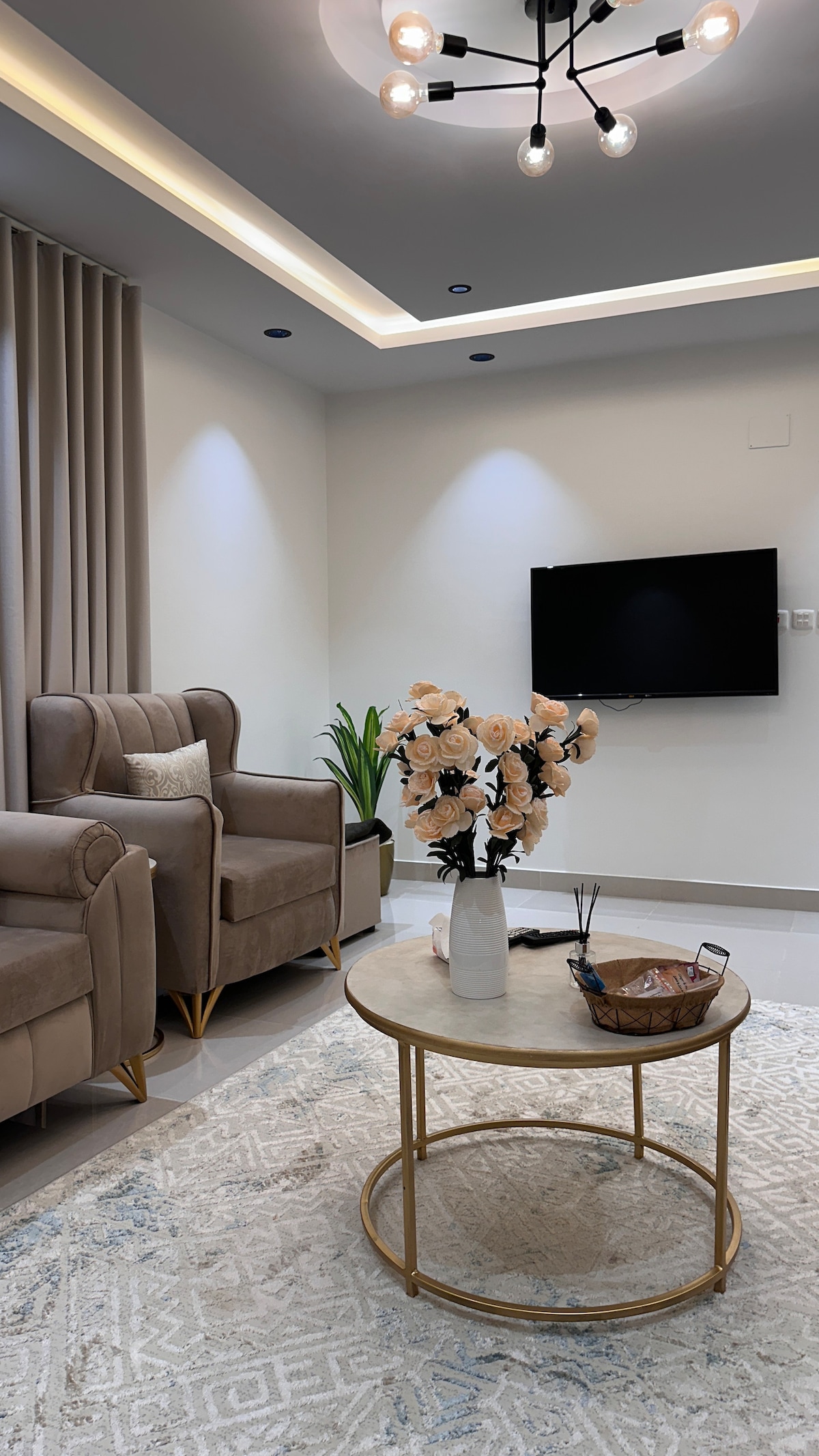 A comfortable seating area features two beige armchairs and a round coffee table. Fresh flowers are displayed in a vase atop the table. A television is mounted on the wall, while subtle lighting highlights the textured rug beneath.